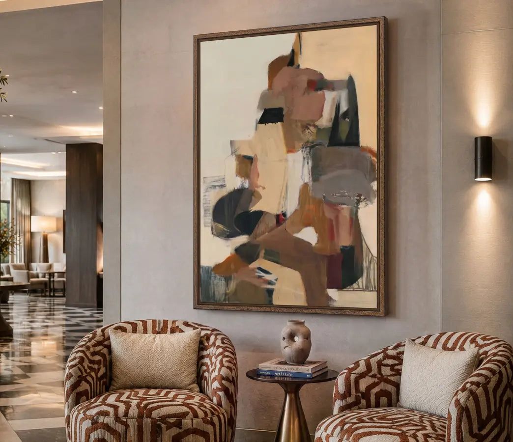 A large framed abstract painting with geometric shapes in beige, brown, gray, and black hangs on a warm beige wall in a modern lobby setting, flanked by two patterned armchairs and a small side table.