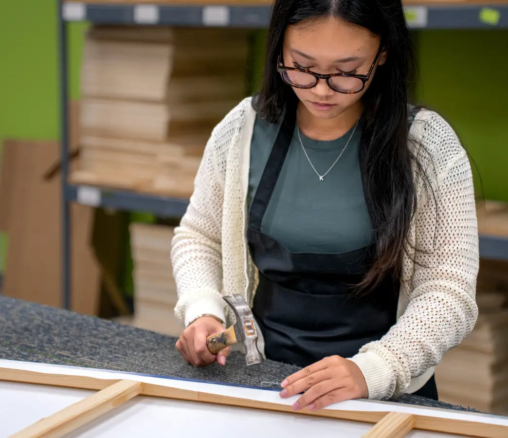 A woman wearing glasses, a cream knit cardigan, and a black apron is shown in a workshop setting, using a hammer to apply adhesive or seal a white sheet attached to a wooden frame.