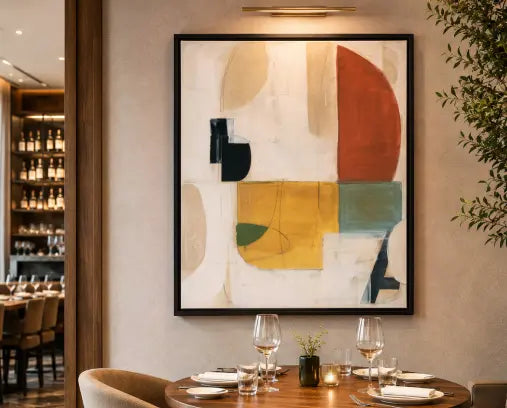 A large framed abstract wall art print hangs above a dining table in a restaurant or cafe. The artwork features geometric blocks of cream, rust red, mustard yellow, sage green, and dark navy on a light background.