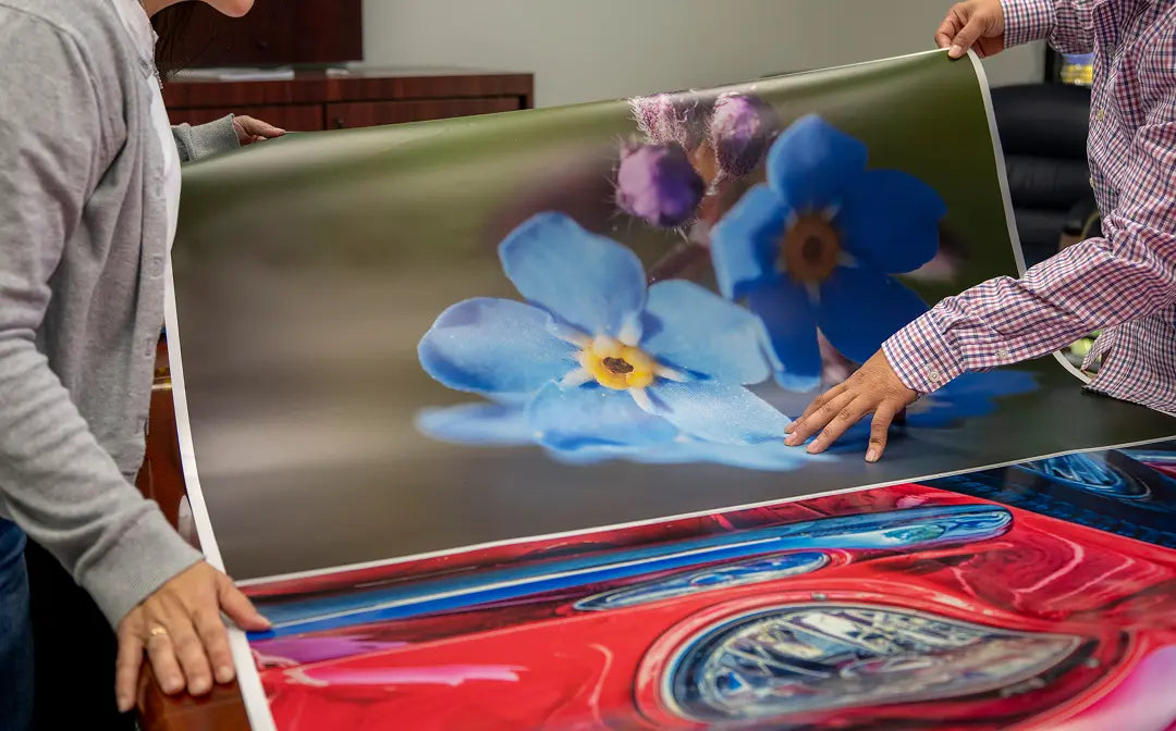 Two people holding up a large printed poster or canvas showing a close-up floral scene with blue forget-me-nots and purple blossoms, while another colorful red and blue artwork is partially visible below.