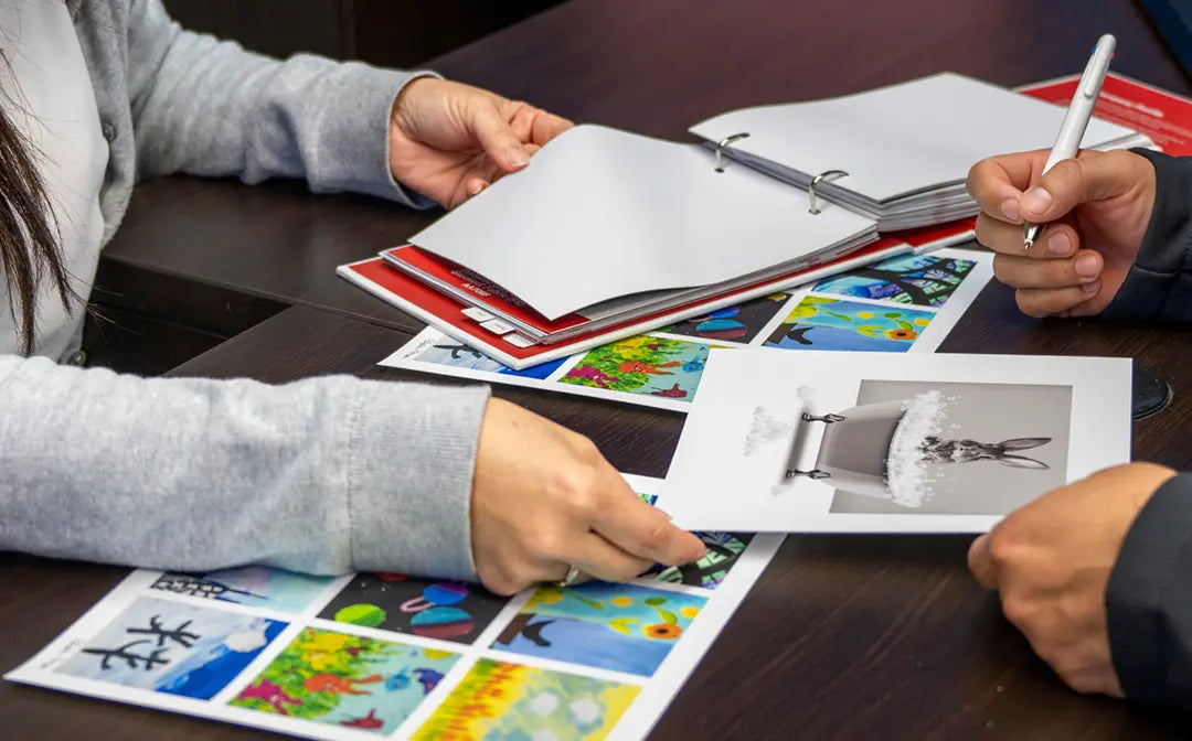 A close-up of a person's hands holding a printed art card over a table with colorful children's paintings and a binder, with another person's hands holding a pen nearby.
