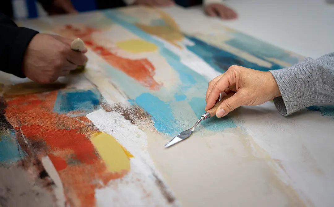 Close-up of a person using a palette knife to apply cream paint onto a large abstract canvas, with other hands visible holding brushes and tools over the artwork.